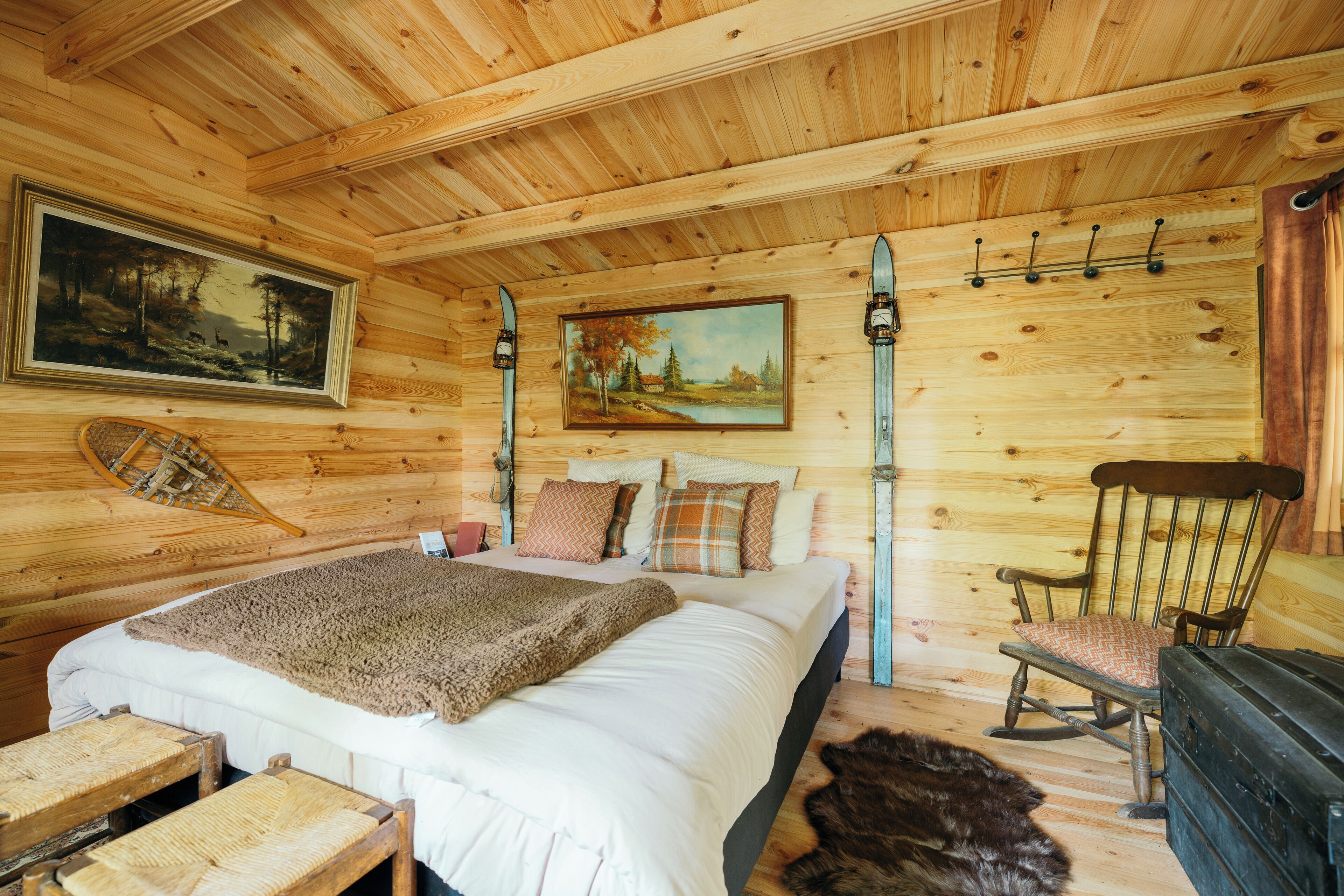 A wooden cabin bedroom with a bed, a chair, a painting, and a ski rack.