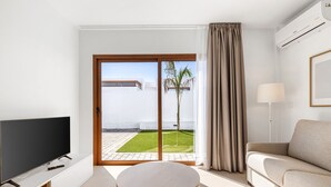 Un salon moderne avec un téléviseur à écran plat, une table basse ronde, un canapé et une porte-fenêtre coulissante donnant sur un balcon avec vue sur un immeuble blanc et un palmier.