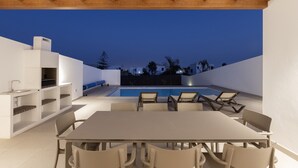 Un espace de restauration extérieur moderne, doté d’une piscine, de poutres en bois au plafond et offrant une vue sur le quartier environnant au crépuscule.