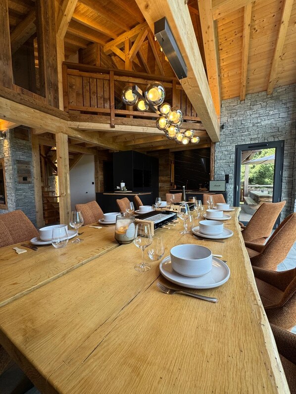 A dining area with a long wooden table set for a meal, surrounded by chairs, and a stone wall in the background.