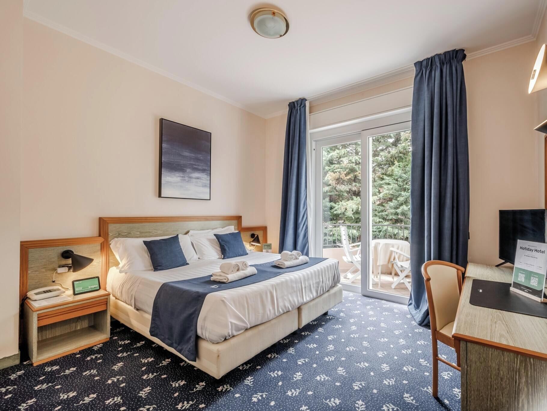 A hotel room with a bed, a desk, a chair, a TV, and a balcony with curtains.