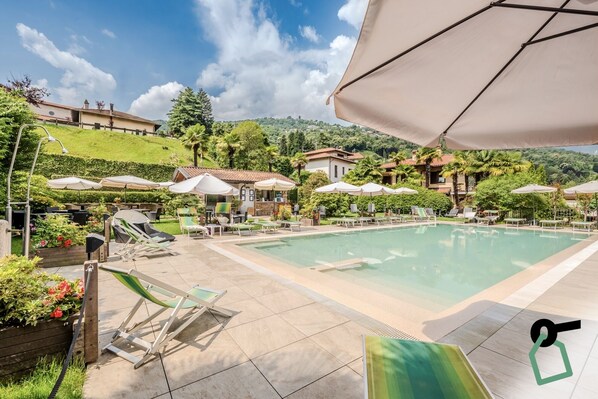 A pool area with lounge chairs and umbrellas, surrounded by greenery and buildings. - Hotiday Room Collection - Stresa (Stresa)