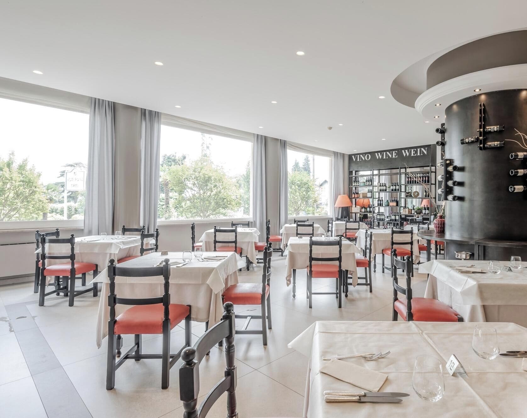 A restaurant with tables and chairs, a wine rack, and a view of trees outside.