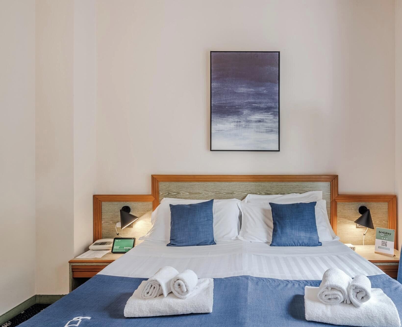 A hotel room with a bed, pillows, a nightstand with a phone and a tablet, and a wall-mounted painting.