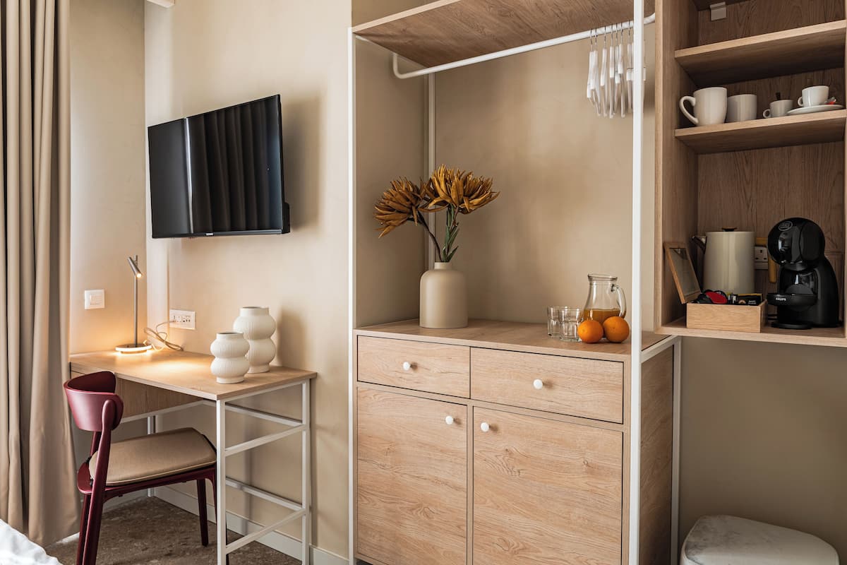 Habitación de hotel con escritorio, silla, televisión, aparador de madera y estantes con tazas y cafetera.