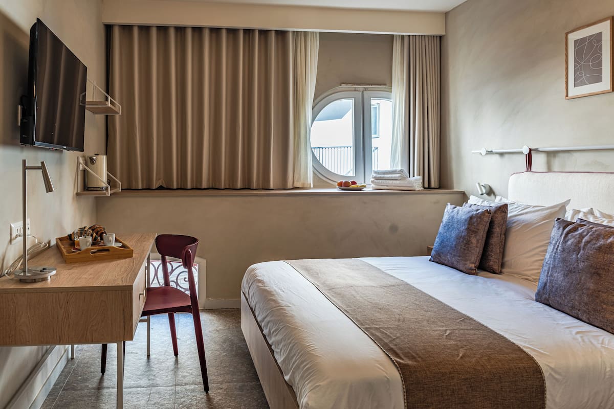 Habitación de hotel con cama, escritorio, silla, televisor, ventana con cortinas y vistas al balcón.