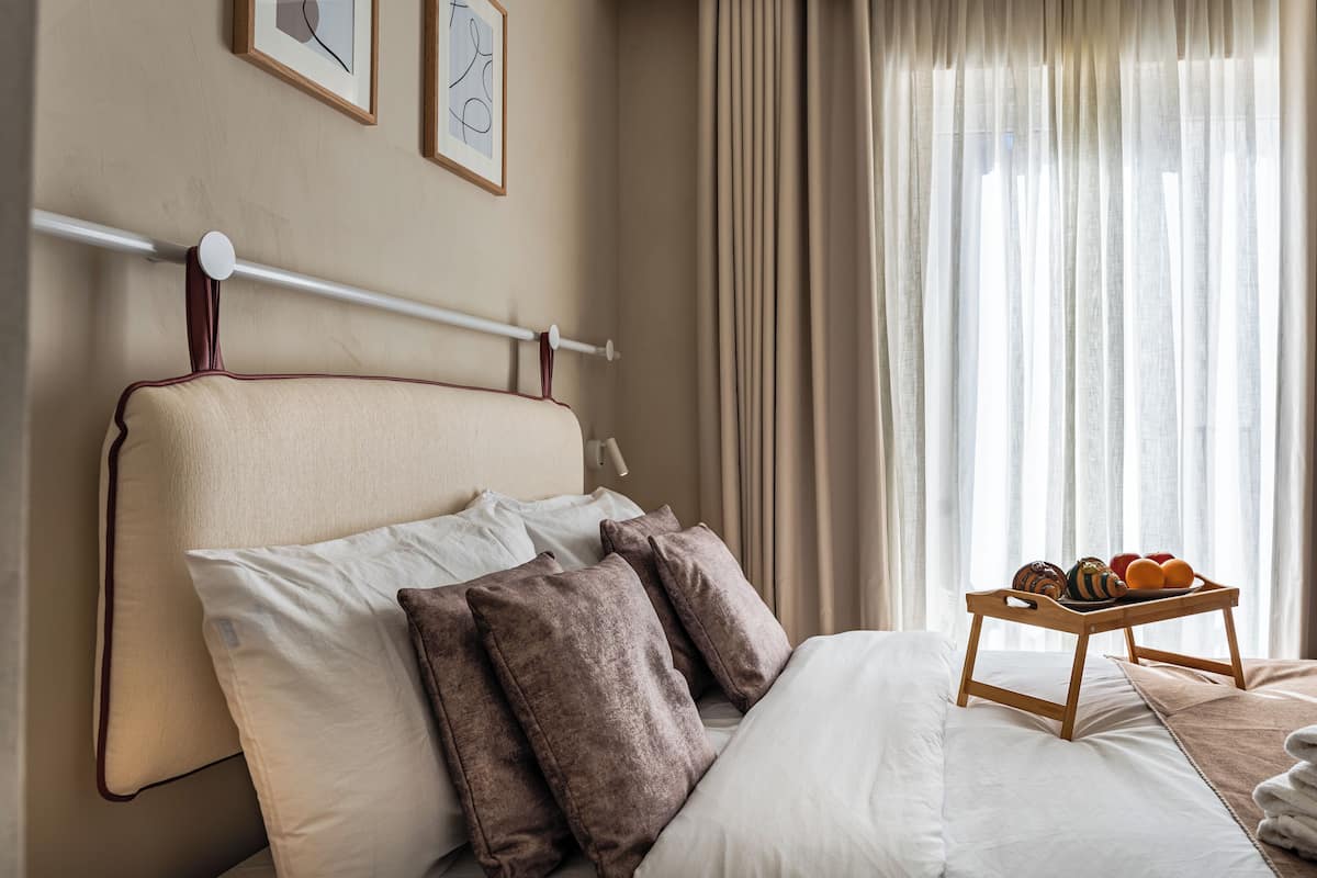 Una cama bien tendida con almohadas, una bandeja con frutas, y vistas a cortinas y una pared con cuadros.
