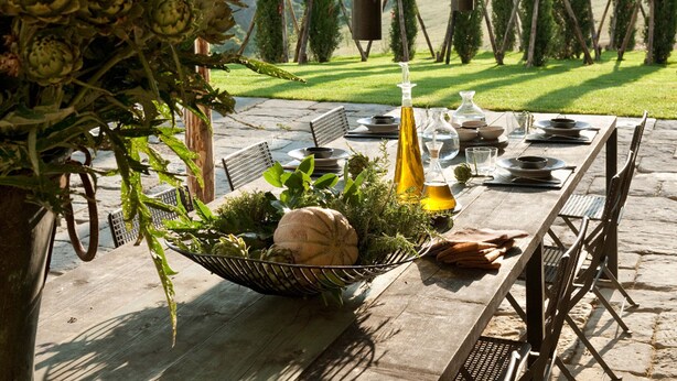 An outdoor dining area with a stone table set for a meal, surrounded by greenery and a view of a grassy landscape with trees. - AMORE RENTALS - Villa Valle with Pool, Garden, Ideal for Weddings (Sarteano)