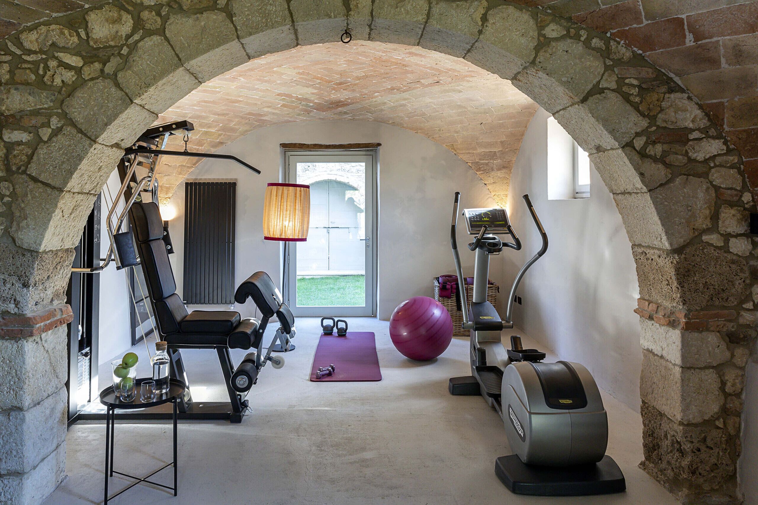 Un arc en pierre mène à une salle de sport privée équipée de matériel d’exercice, d’un tapis de yoga et d’un grand ballon de gymnastique.