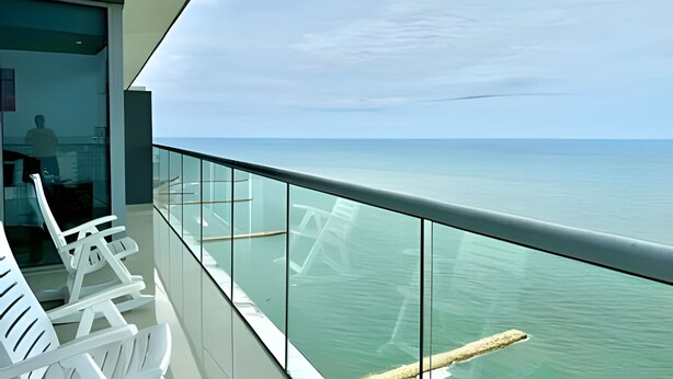 Un balcon avec vue sur la mer, des garde-corps en verre et des chaises blanches.