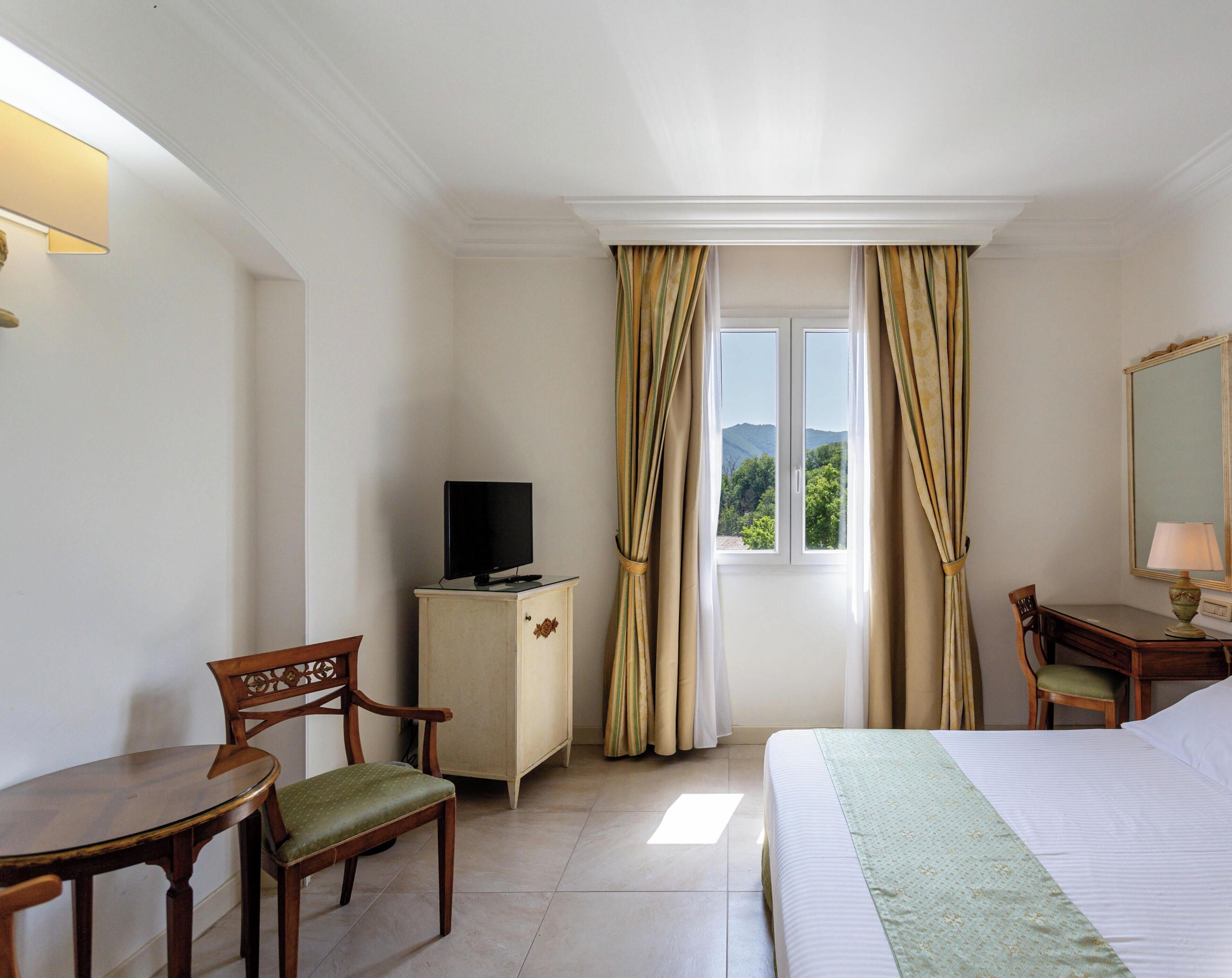 Habitación de hotel con una cama, un escritorio con silla, un televisor sobre un mueble bajo y una ventana con cortinas.