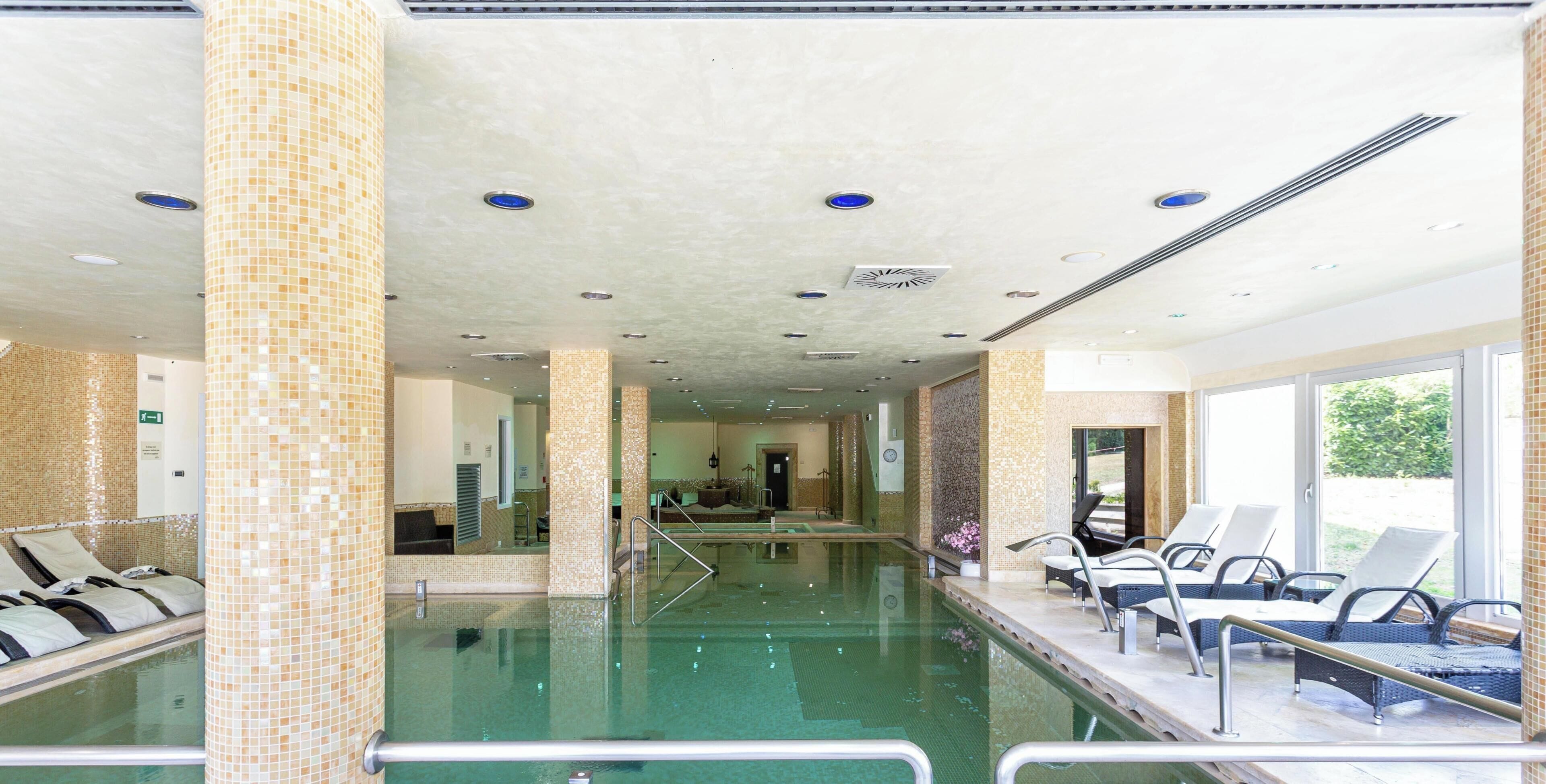 Una piscina interna con pareti e soffitto piastrellati, sedie a sdraio e vista libera sull'esterno.