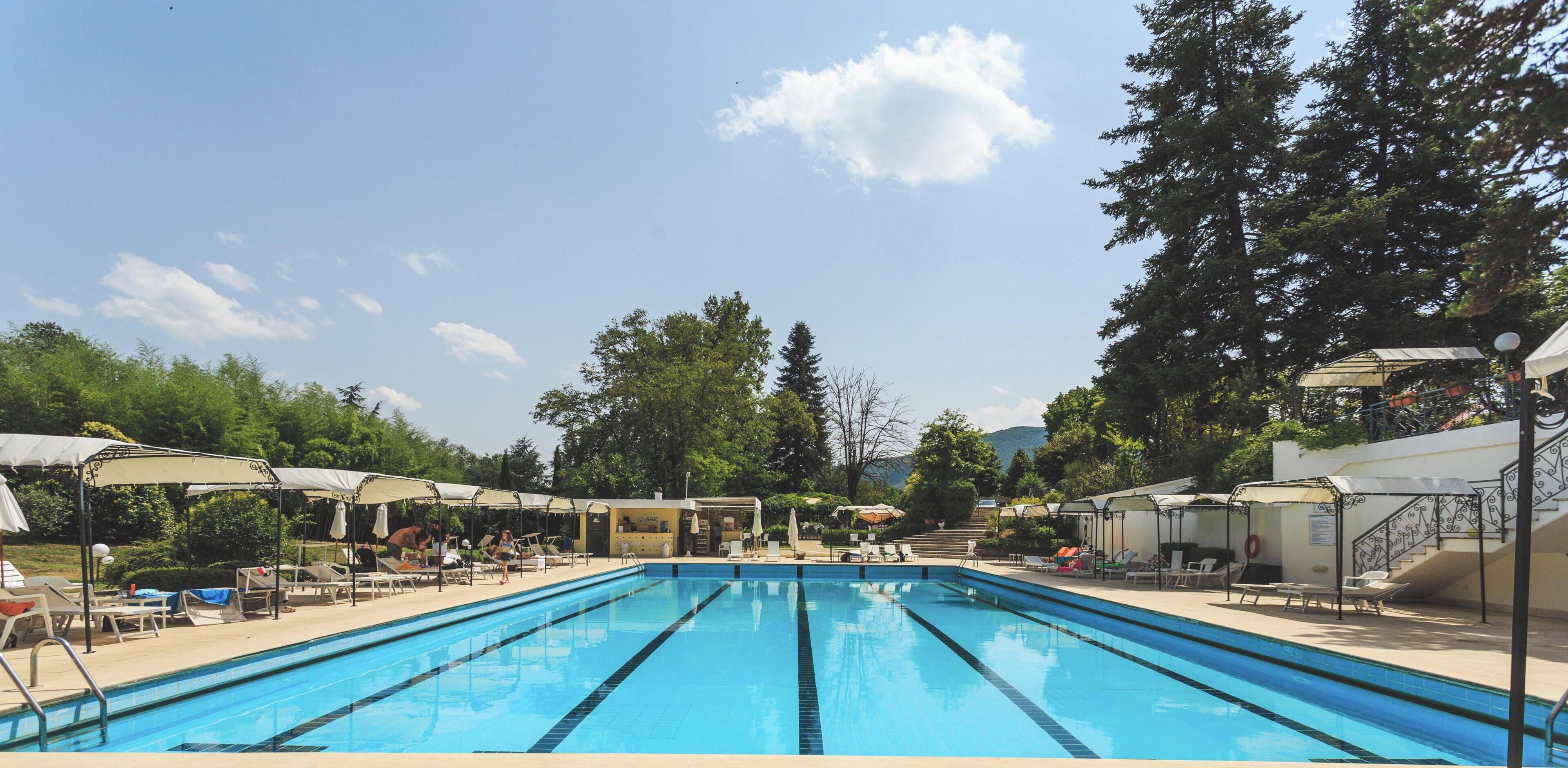 Una piscina con sillas de descanso y sombrillas, rodeada de árboles y un cielo despejado.