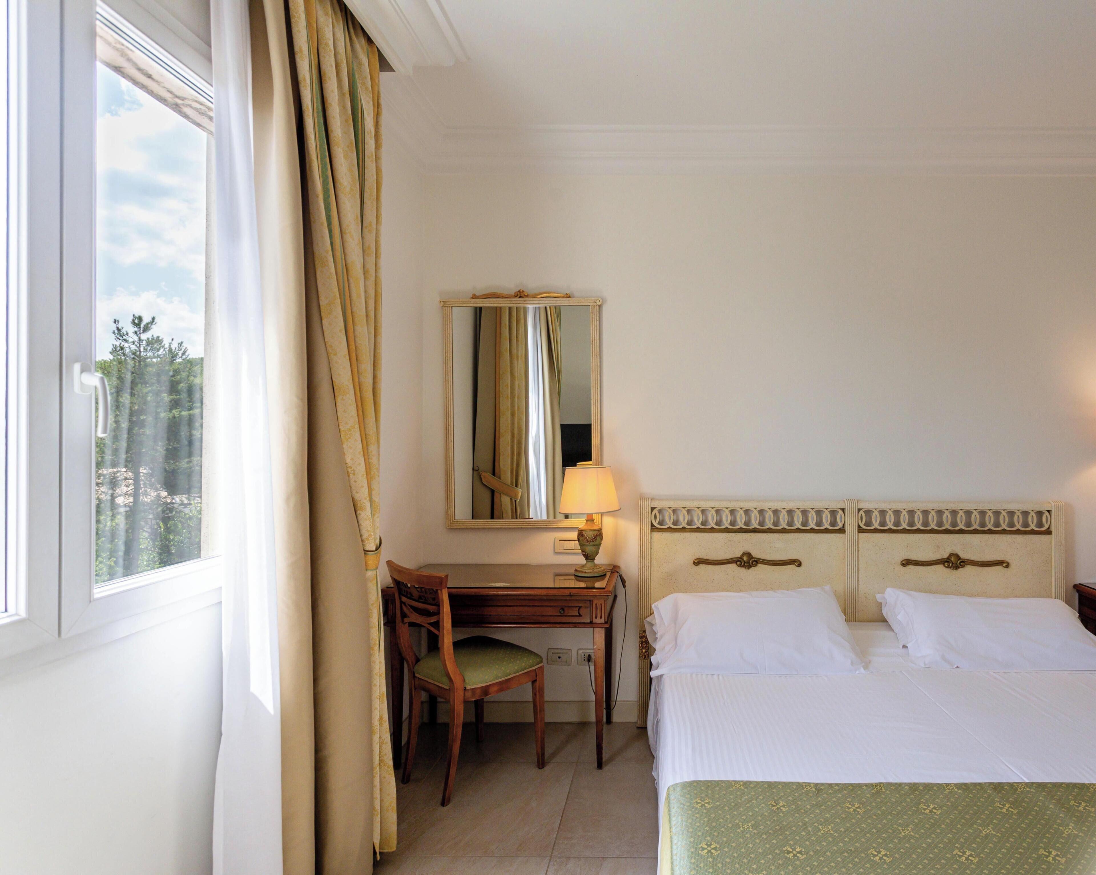 Ein Hotelzimmer mit Bett, Schreibtisch, Stuhl, Lampe und einem fenster mit Vorhängen.