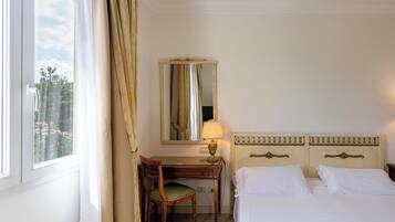 Ein Hotelzimmer mit Bett, Schreibtisch, Stuhl, Lampe und einem fenster mit Vorhängen.