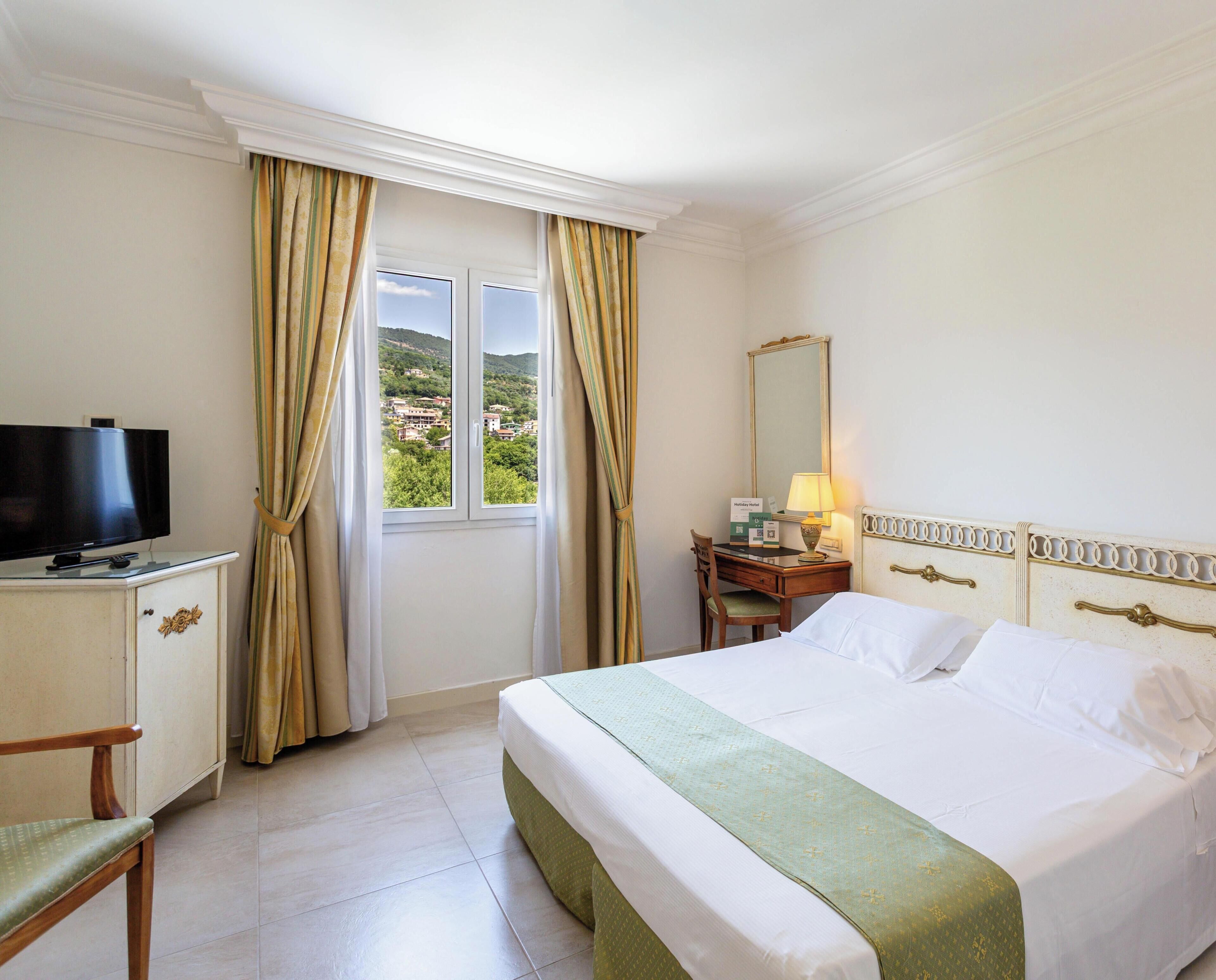 A hotel room with a bed, a desk, a chair, a television, and a window with curtains.