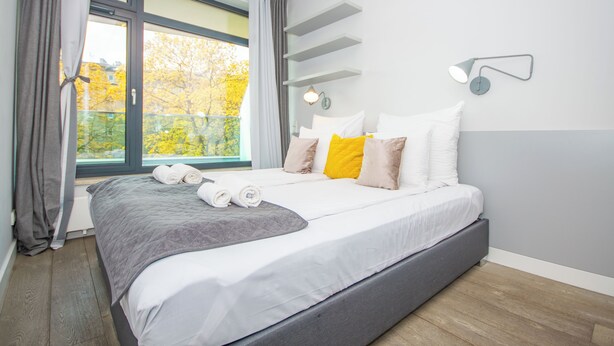 A modern bedroom with a large bed, a window with curtains, a bedside table, and a wall-mounted lamp. - 1-bedroom in city center | balcony | close to the Main Square and Jewish Quarter (Kraków)