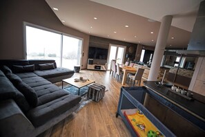 A modern living room with a large sofa, a dining area with chairs, a kitchen with a bar, and a view through large windows.