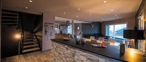 A modern kitchen with a stone island, a dining area with chairs, a staircase, and a large window.