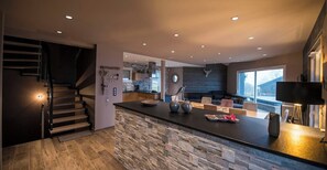 A modern kitchen with a stone island, a dining area with chairs, a staircase, and a large window.