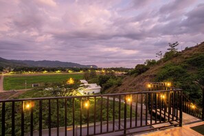 Uma varanda com guarda-corpo de metal e luzes de Natal, com vista para uma paisagem verde com um edifício branco e uma estrada.