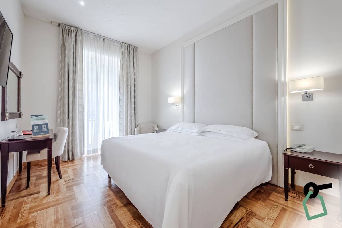 A hotel room with a large bed, a desk with a chair, a telephone, and a wall-mounted lamp.