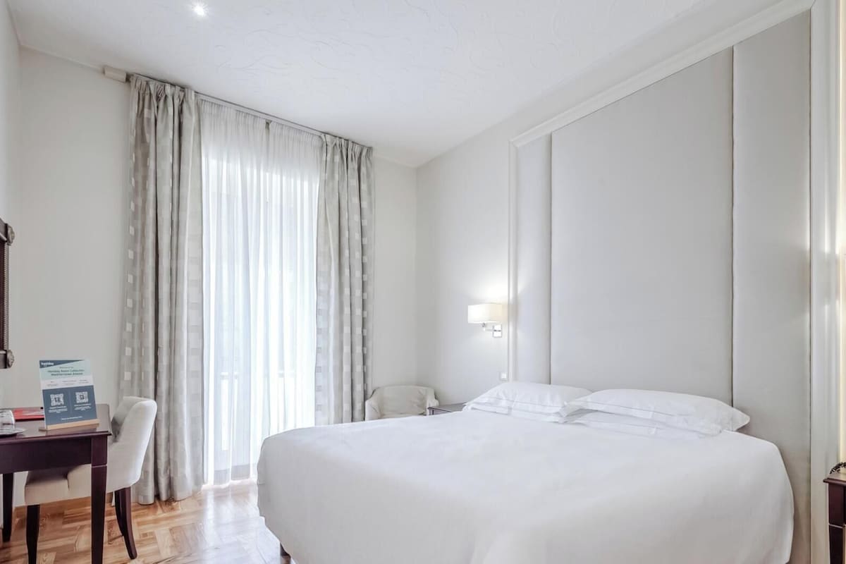 A hotel room with a bed, a desk, a chair, a lamp, and a telephone.