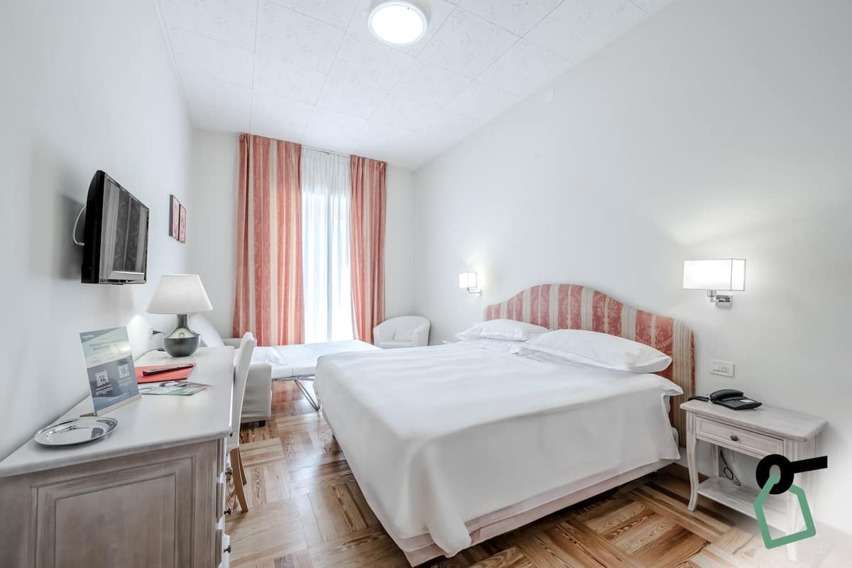 A hotel room with a large bed, a desk with a lamp, a chair, a TV mounted on the wall, and a window with curtains.