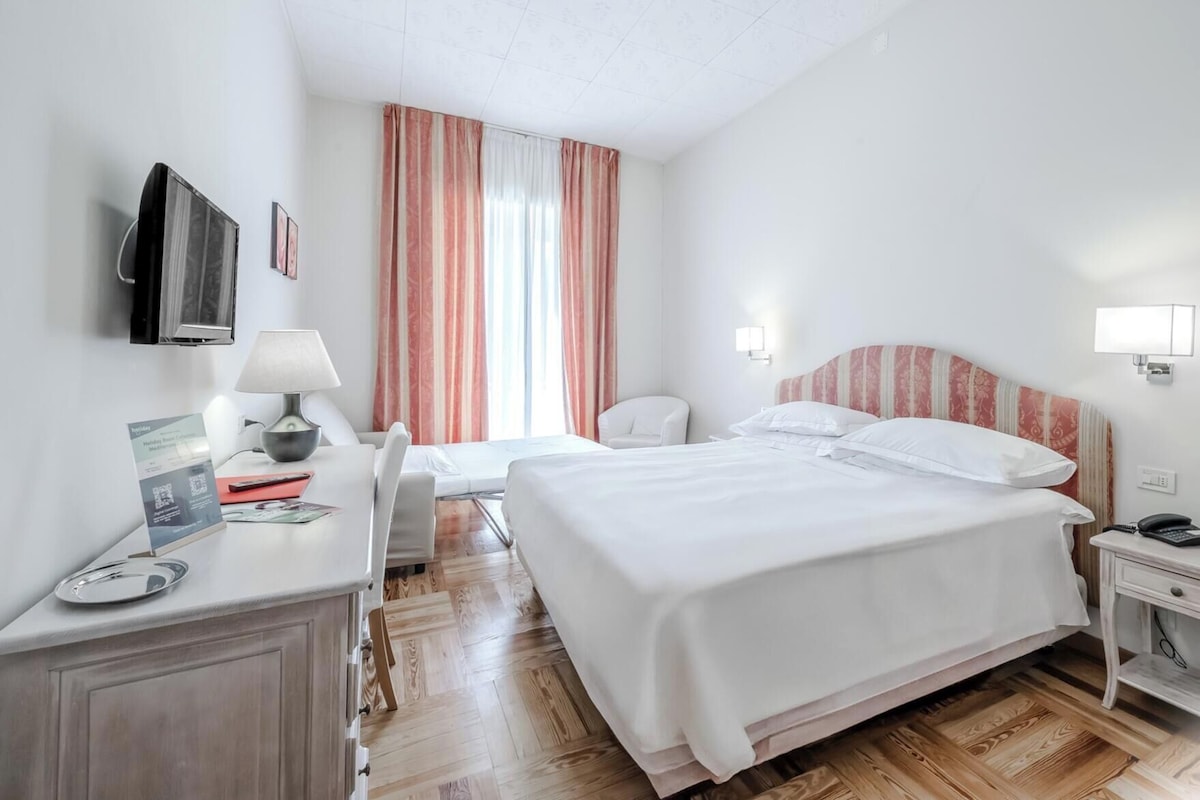 A hotel room with a bed, bedside tables, a desk with a lamp, a television, and a window with curtains.