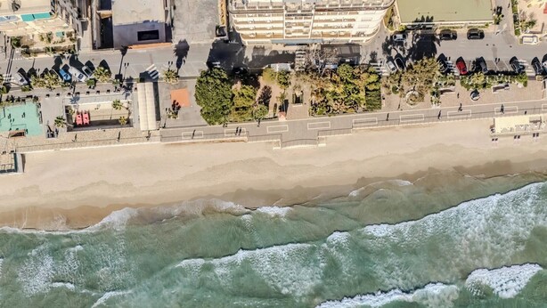 Vista aérea de una zona costera con playa de arena, paseo marítimo arbolado y un complejo de edificios al fondo.