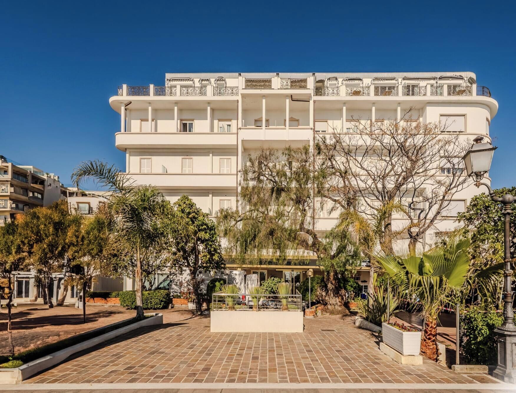 Un hotel a più piani con un cortile centrale, circondato da alberi e un vialetto lastricato.