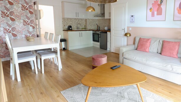 A modern kitchen with white cabinets, a dining area with a white table and chairs, a beige sofa with pink cushions, and a wooden coffee table. - 123home- Garden & spa (Serris)