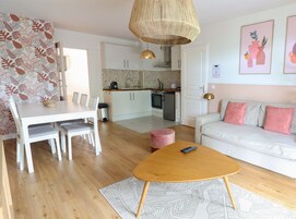 Una cocina moderna con armarios blancos, un comedor con una mesa y sillas blancas, un sofá color beige con cojines rosados y una mesa de centro de madera.