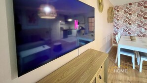 A modern living room with a large flat-screen TV mounted on the wall, a wooden coffee table, a dining table with chairs, and a patterned wallpaper. - 123home- Garden & spa (Serris)