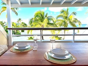 A dining table set with plates, bowls, and glasses, overlooking a beach with palm trees.