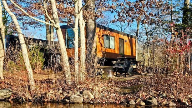 Una cabaña junto a un lago, con fachada de madera, un balcón y una pequeña zona de estar al aire libre.