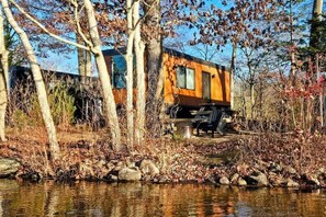 Una cabaña junto a un lago, con fachada de madera, un balcón y una pequeña zona de estar al aire libre.