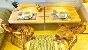 Un comedor con dos mesas de madera dispuestas para dos comensales, cada una con platos, tazas y cubiertos blancos. Hay dos sillas tejidas y una pequeña planta en maceta a la izquierda.