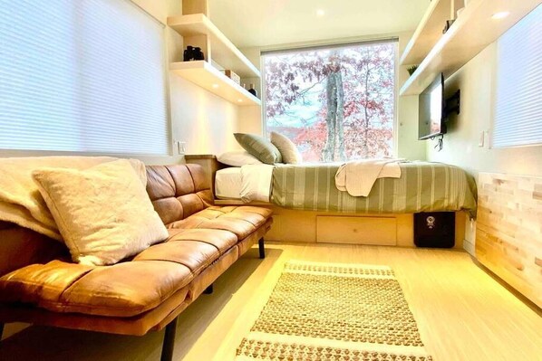 Un dormitorio moderno con sofá, cama, un ventanal con vistas a árboles, estanterías empotradas y un televisor de pantalla plana.