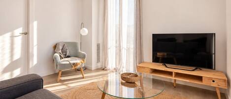 Una sala de estar moderna con un sofá, una mesa de centro de vidrio, un televisor sobre un mueble de madera, una silla con una manta y un ventanal con cortinas translúcidas.
