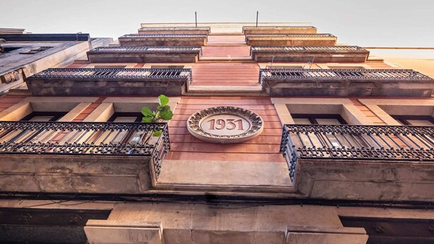 Un edificio con una placa destacada que dice '1931', varios balcones con barandillas de metal y una entrada central con escalones.
