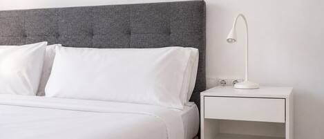 A neatly made bed with white linens, a grey upholstered headboard, and a white bedside table with a lamp.