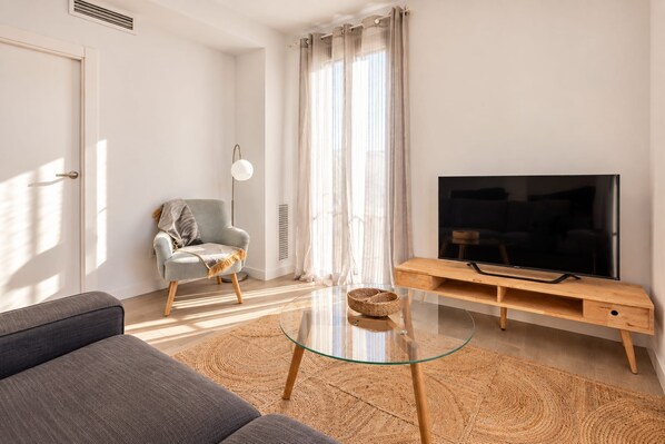 Un salon moderne avec un canapé, une table basse en verre, un téléviseur posé sur un meuble en bois, un fauteuil avec un plaid et une grande fenêtre ornée de rideaux légers.