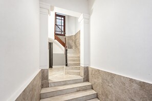 Un escalier avec des marches en marbre menant à un palier, bordé de murs blancs et d’une porte en bois.