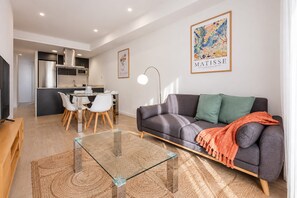 Una sala de estar moderna con una mesa de centro de vidrio, un sofá gris y un comedor con sillas blancas.