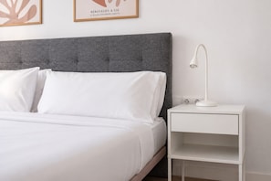 Una cama bien hecha con sábanas blancas, un cabecero tapizado de gris y una mesita de noche blanca con una lámpara.