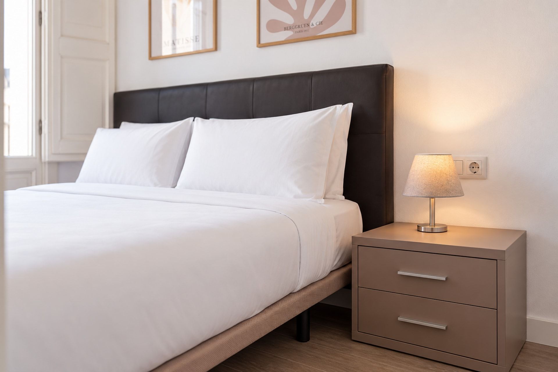 A neatly made bed with white linens, a dark headboard, and a bedside table with a lamp and drawers.