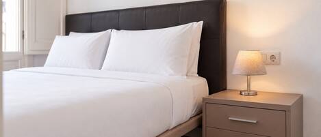 A neatly made bed with white linens, a dark headboard, and a bedside table with a lamp and drawers.