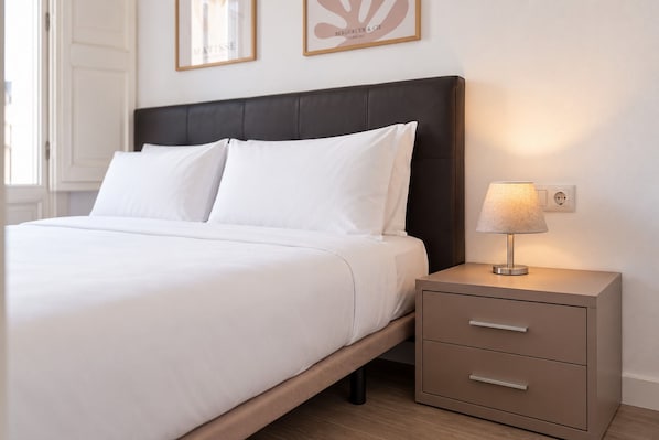 A neatly made bed with white linens, a dark headboard, and a bedside table with a lamp and drawers.