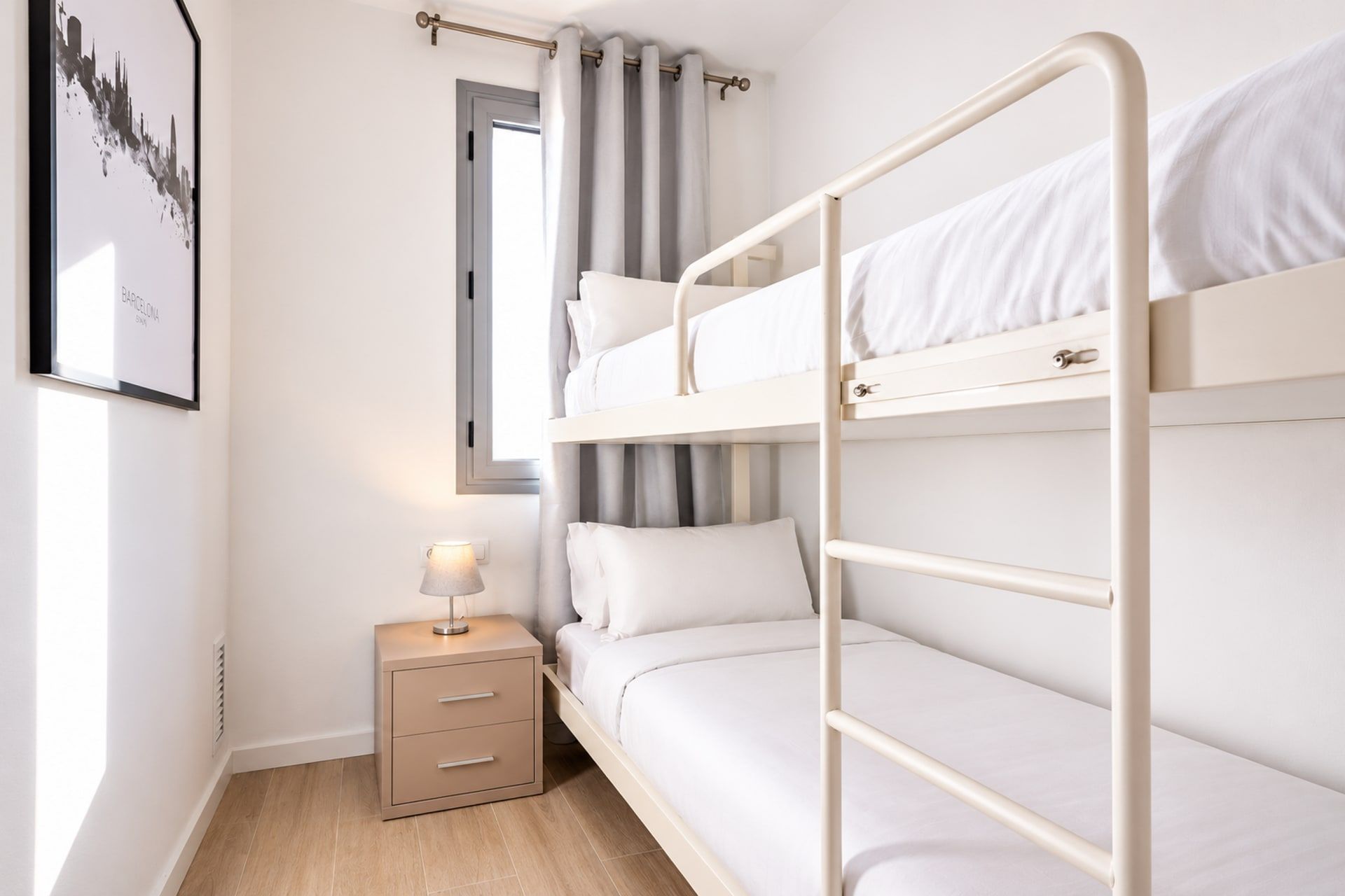 Una cama individual superpuesta con una mesita de noche y una lámpara, una ventana con cortinas, y un cuadro en la pared.